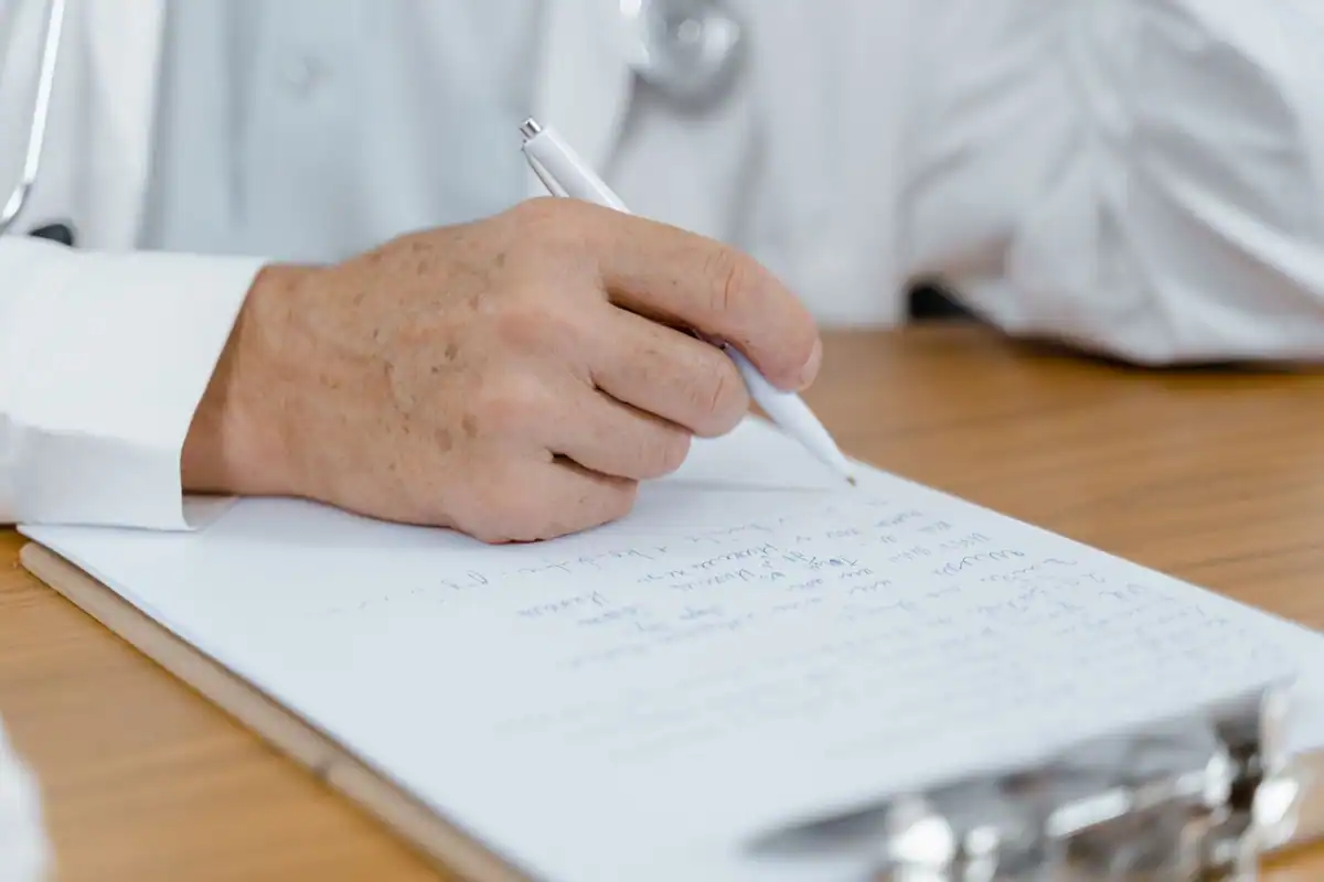 Mâna unui medic completează cu pixul un document pe un birou de cabinet
