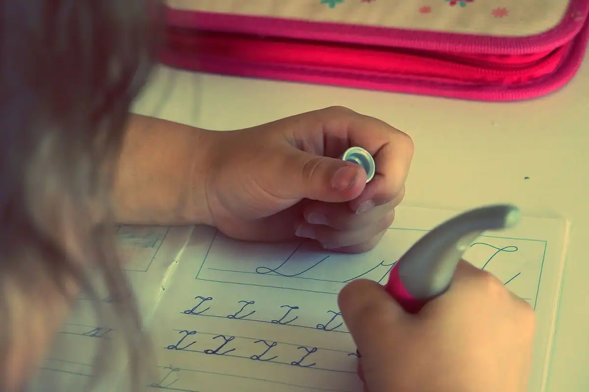 Copil care scrie litere cu un pix pe hârtie, la teme, într-un cadru școlar