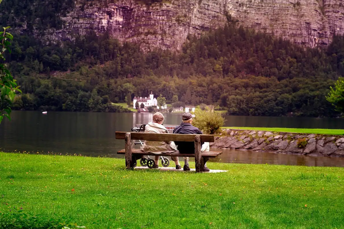 Doi vârstnici stau pe o bancă în parc, lângă un lac