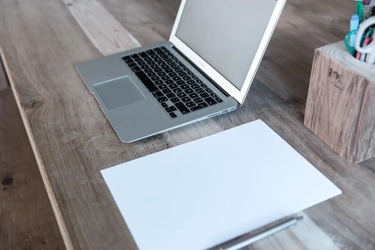 Laptop, foi de hârtie și un pix pe o masă de lemn, într-un spațiu de lucru