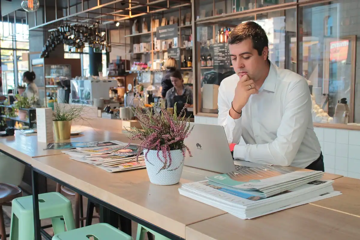 Bărbat lucrează concentrat la laptop într-o cafenea cu mese lungi