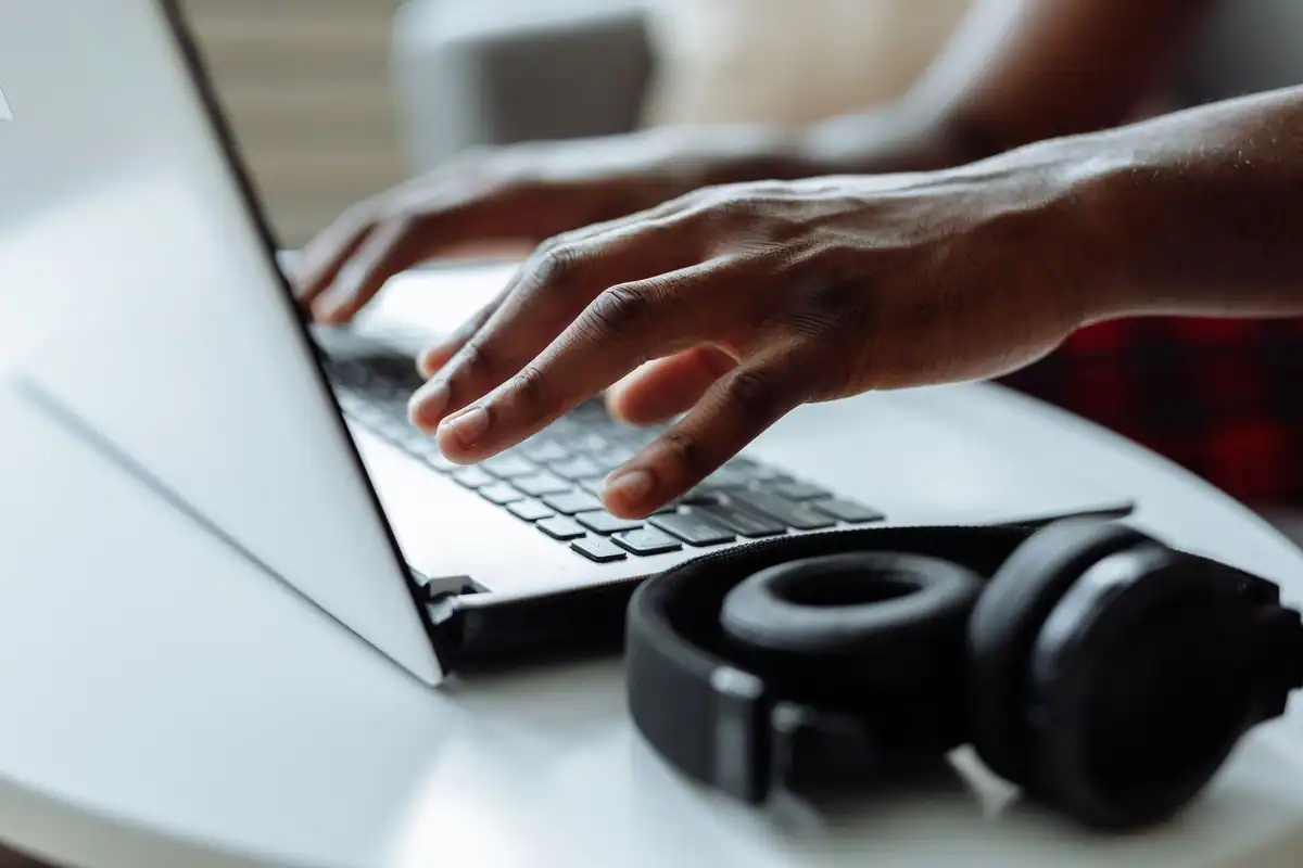 Persoană cu căști care scrie la laptop, așezată la o masă, într-un decor de lucru la distanță