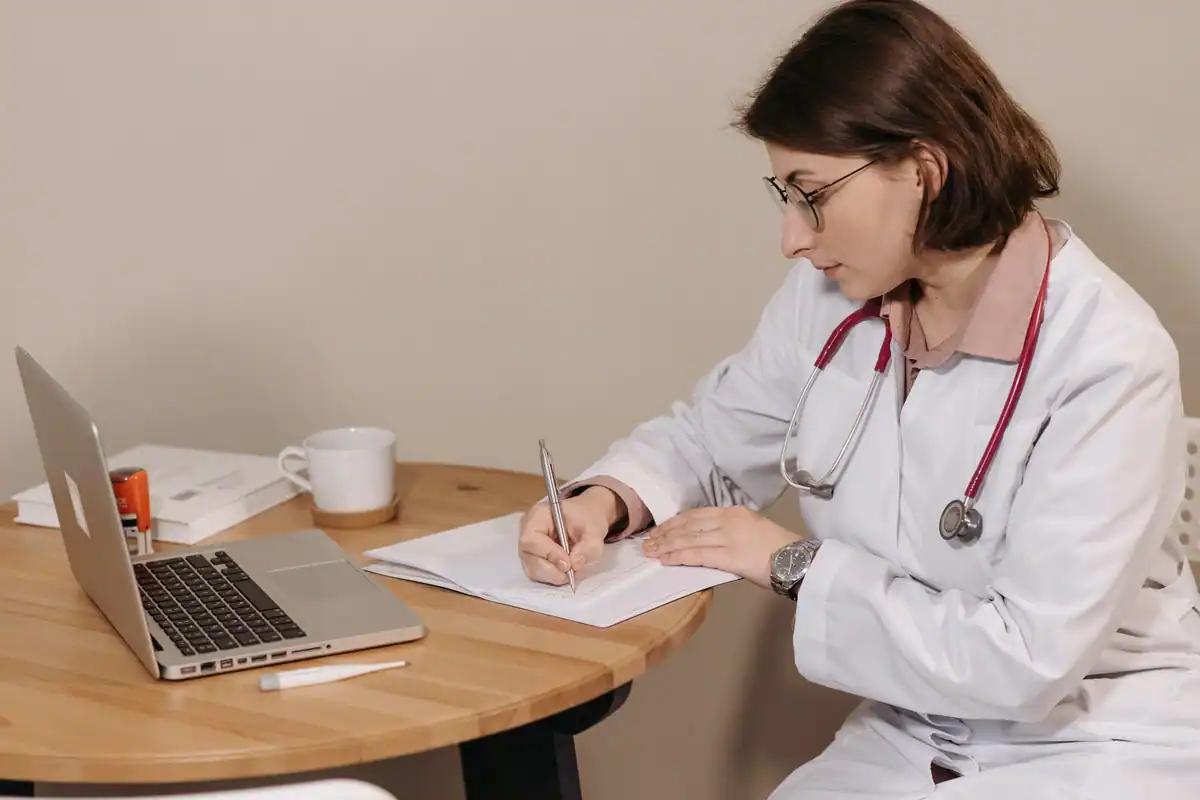 Femeie medic cu stetoscop care scrie la birou, lângă un laptop și niște notițe