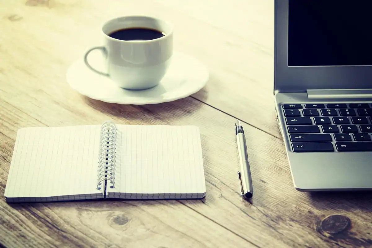 Laptop, caiet, pix și cană de cafea pe o masă de lemn, într-un spațiu de lucru de acasă